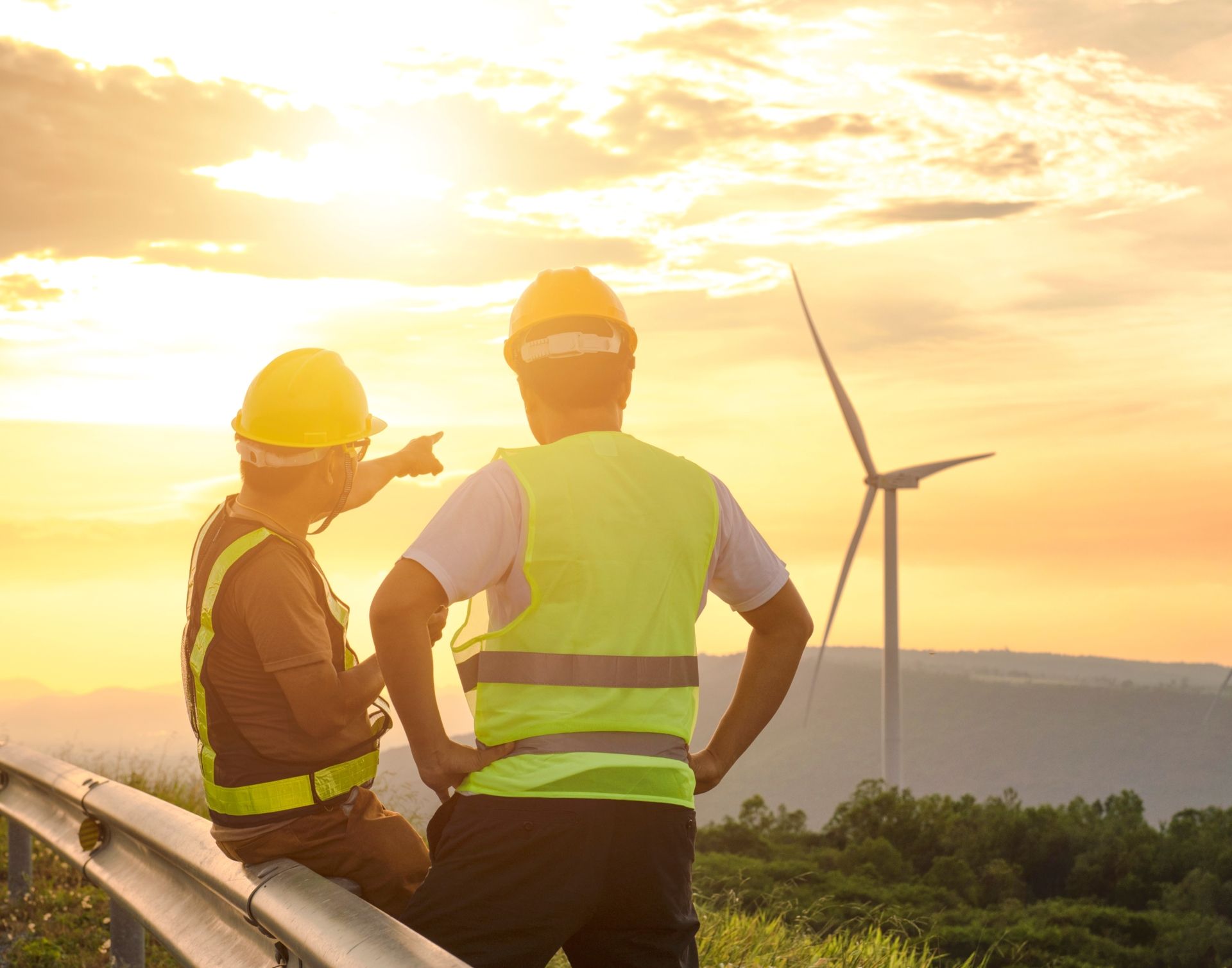 Doi bărbați în veste și căști de protecție inspectează turbinele eoliene în aer liber.