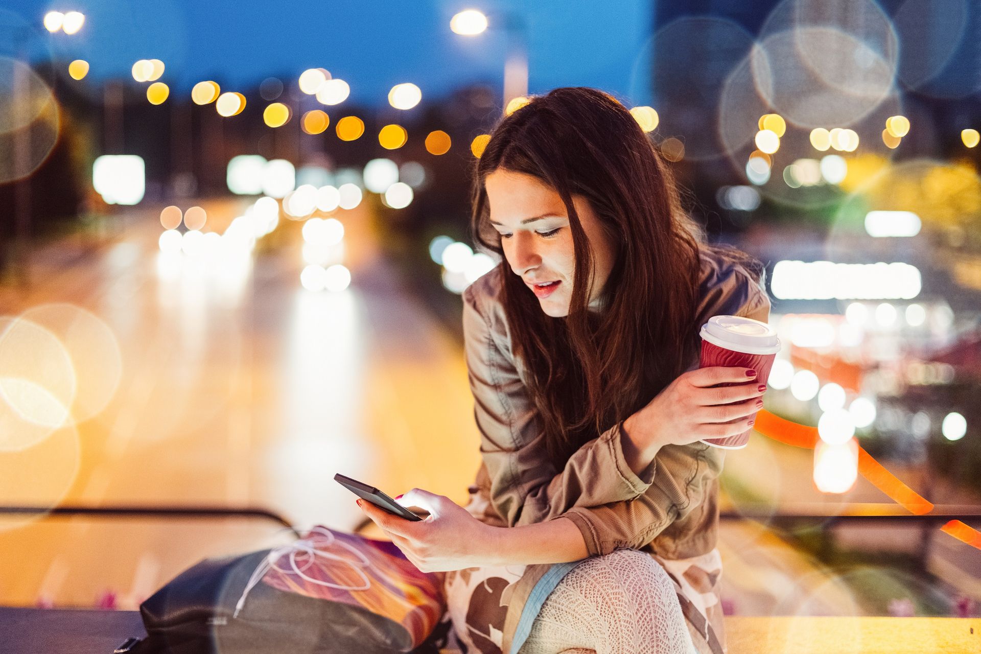 Femeie stând pe o bancă, ținând un telefon mobil și o ceașcă de cafea, zâmbind într-un mediu urban.